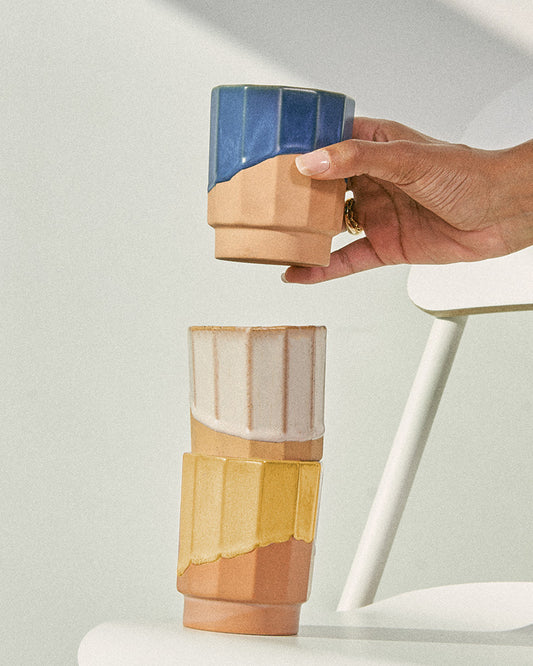 Foxglove 8 oz. Stackable Stoneware Tumbler (In-use): Hand places blue-glazed tumbler atop two stacked terracotta tumblers, white and yellow glazed