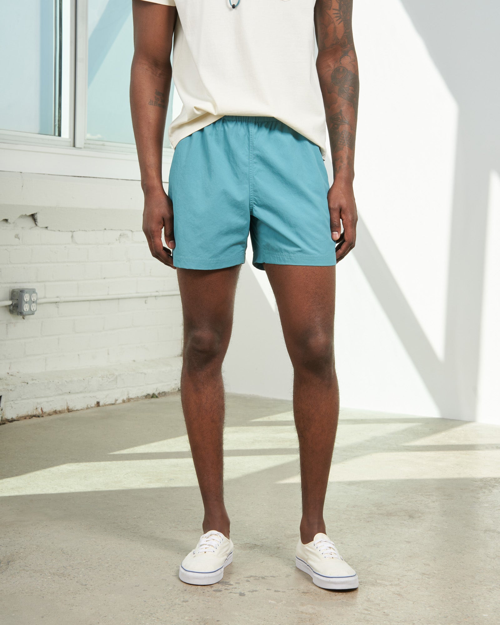 Teal Recycled Sport Short (In-use): Man wearing teal elastic-waist shorts with white sneakers outdoors