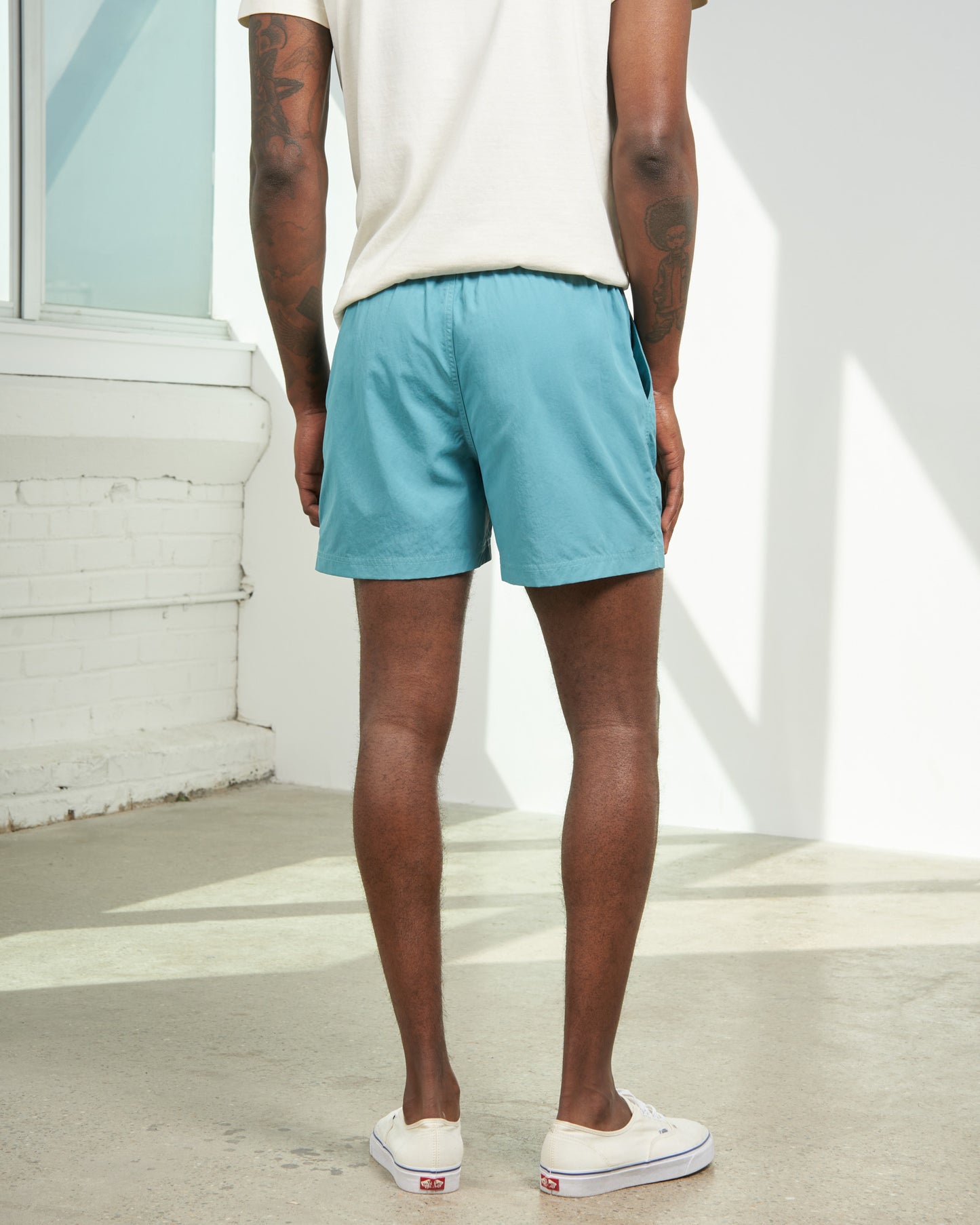 Teal Recycled Sport Short (Back): Man wearing teal elastic-waist shorts, cream tee, white Vans sneakers