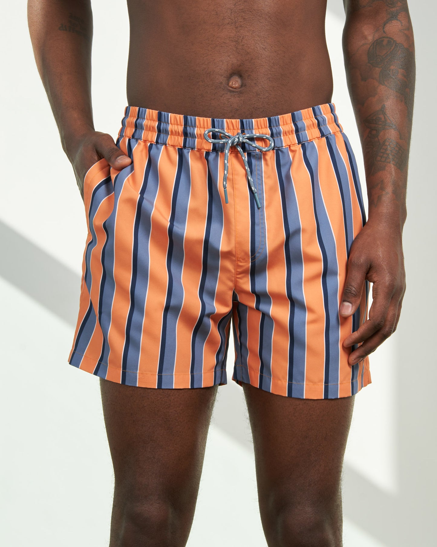 Orange Ochre Recycled Swim Trunk - 5" (In-use): Orange and blue vertical striped shorts with drawstring waist on model