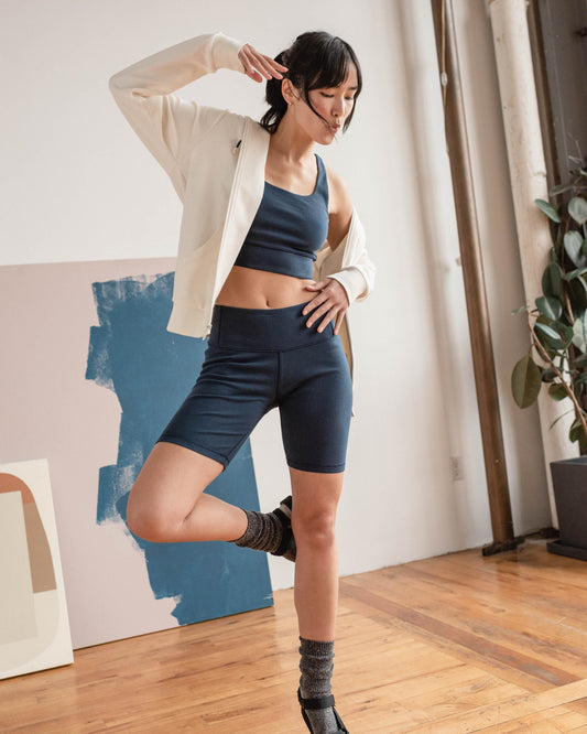 Midnight EcoKnit™ Ribbed Bike Short (In-use): Woman balancing on one leg wearing navy ribbed bike shorts and cream jacket