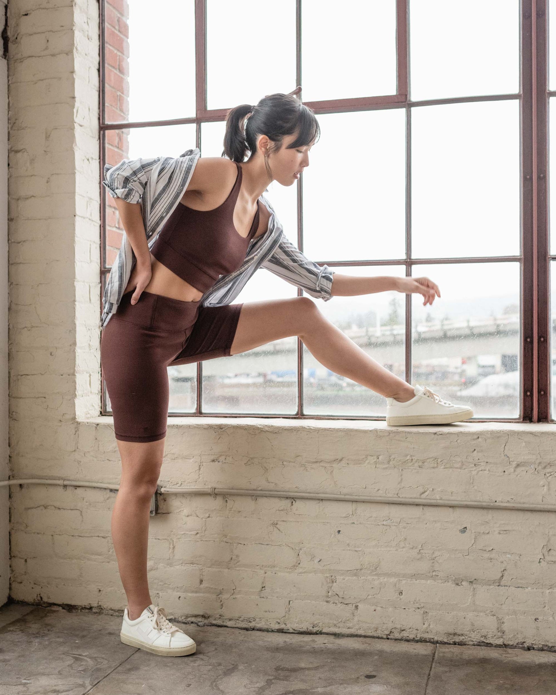 Chocolate EcoKnit™ Ribbed Bike Short (In-use): Model in dark brown ribbed bike shorts raises leg onto windowsill indoors